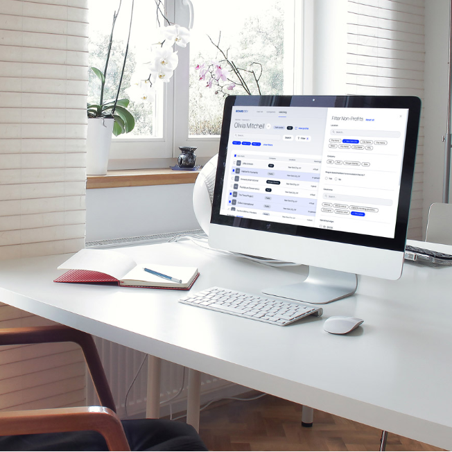 A bright, modern office featuring a desktop iMac computer on a white desk, displaying a business or workflow management dashboard interface, with a keyboard, mouse, plant, and open notebook nearby.