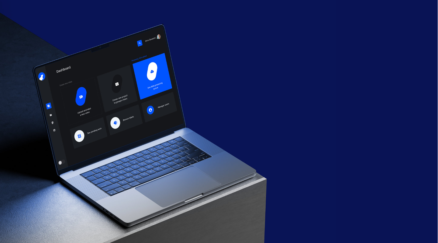 A laptop on a dark surface with a deep blue background, displaying a modern dark-themed dashboard interface featuring large blue and white icons for uploading and labeling videos, seeing pending work, managing users, and browsing assets.