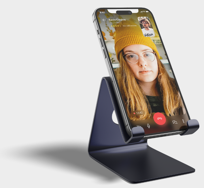 A smartphone on a sleek, black stand displaying a video call with a woman wearing glasses and a yellow beanie.