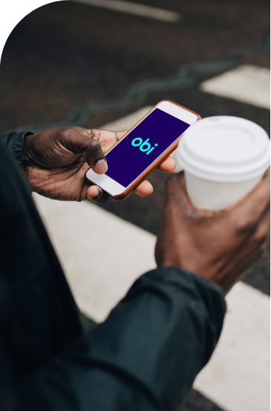 A person standing outside, holding a smartphone showing the "obi" logo on the screen in one hand and a white disposable coffee cup in the other.